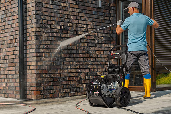 A man wearing a blue shirt, gray pants, and yellow boots uses a black and gray high-pressure water washer to clean a brick wall outdoors.